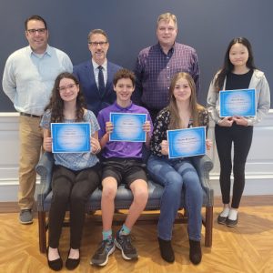 Three student winners sit on a couch smiling at the camera. Another student stands behind the couch with an advisor, the Secretary of Education and a DDOE staff member, smiling at the camera.