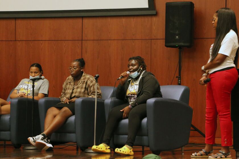 A girl wearing black clothes and with a face mask pulled over her chin speaks into a microphone while seated as two other people sit and listen and a woman in red pants looks on.