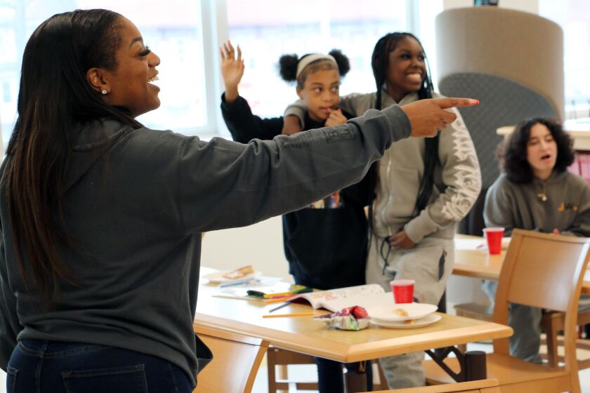 A woman in a dark long sleeve shirt and jeans points and laughs in a room. Three school age children are in the background.