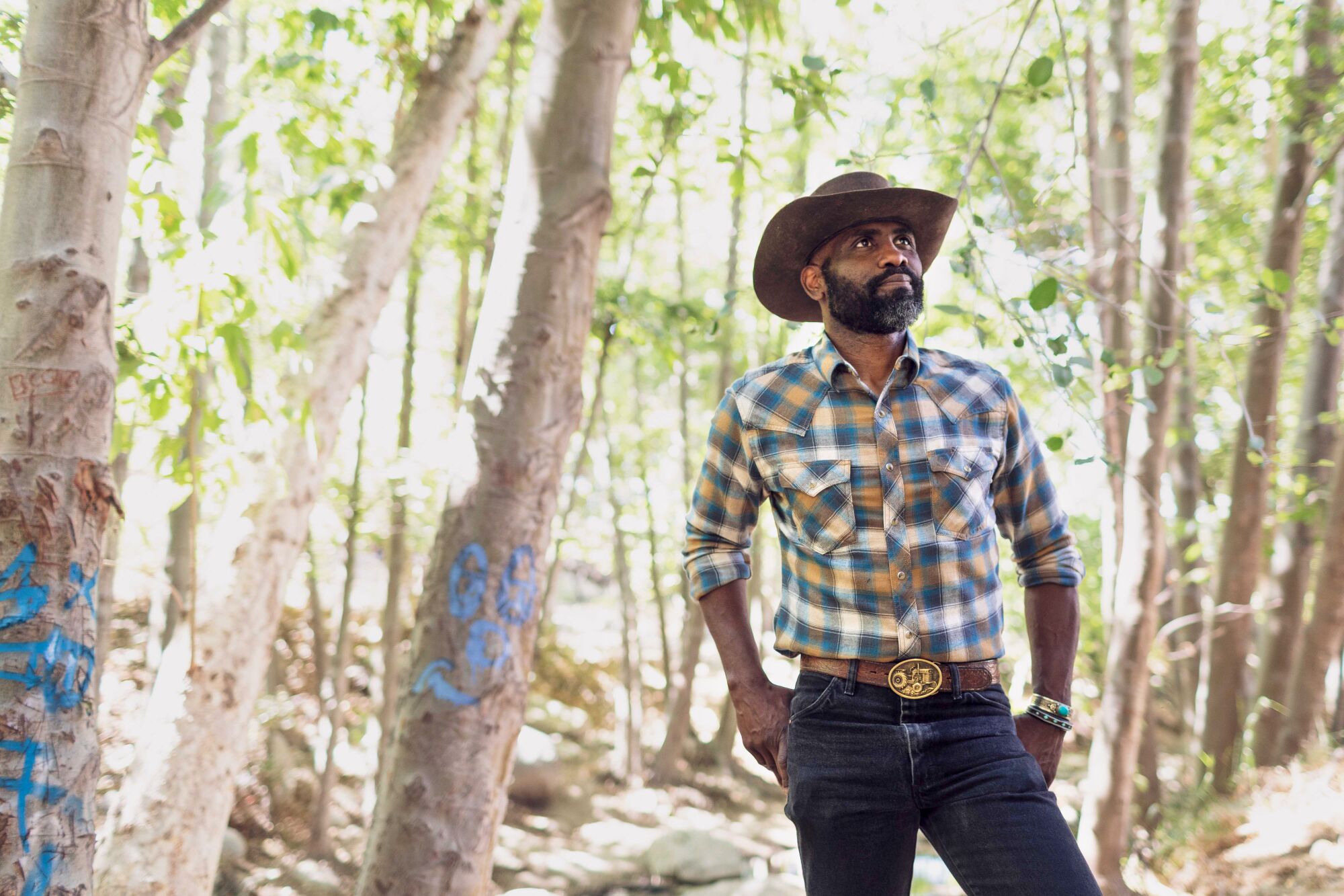 Hop Hopkins, wearing a checkered shirt, jeans, and a brimmed hat, stands in front of leafy trees