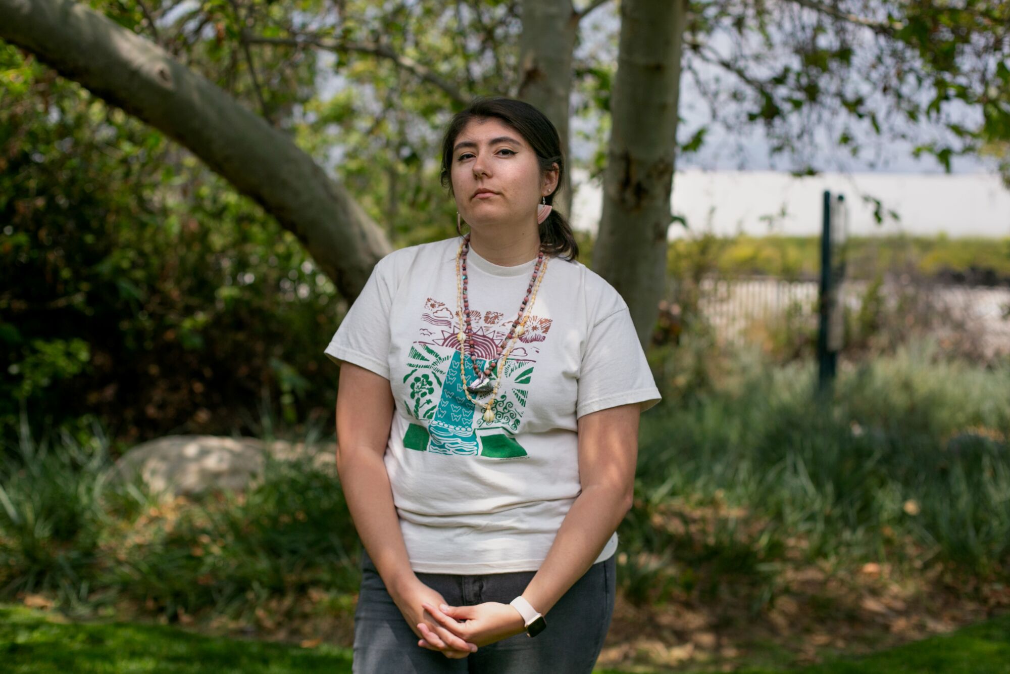 A young woman with dark hair and wearing a T-shirt stands in a shade of a tree behind her. 