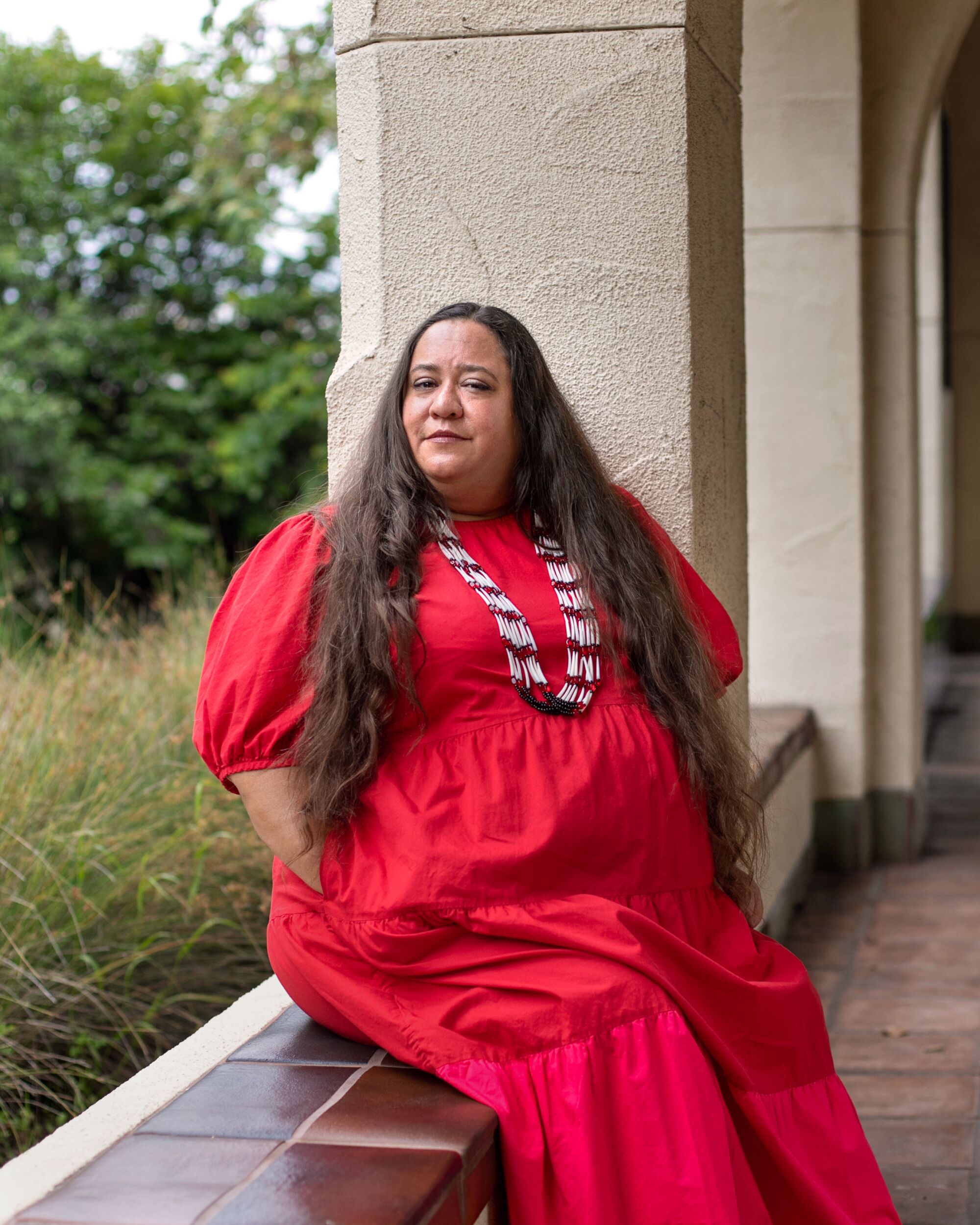 Angela Mooney D'Arcy, wearing a long red dress, sits on a side of an archway.