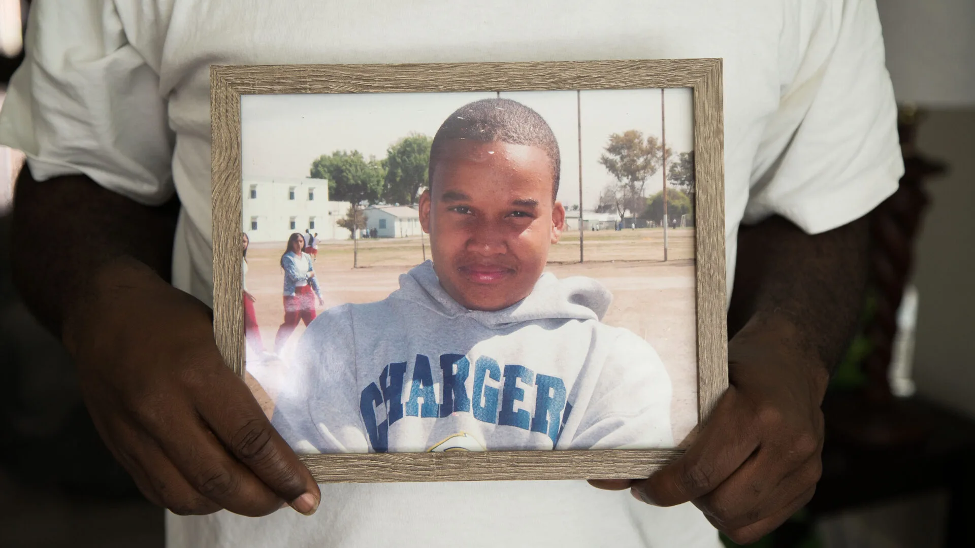 A Black man's hands holds a framed photo of Avery Cody Jr., a teenage Black boy. Avery is wearing a grey hoodie that says CHARGERS in blue font. His hair is cropped short, and he smiles without teeth. 