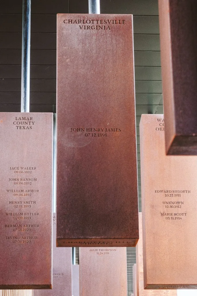 A close-up view of a series of monuments in a memorial. Focused in the center of the photo, a rust-colored metal box hanging from a ceiling. It reads 