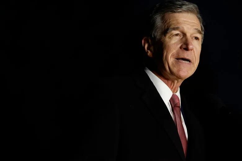 Roy Cooper speaks, wearing a black blazer, red tie and white shirt. The background is very dark. 