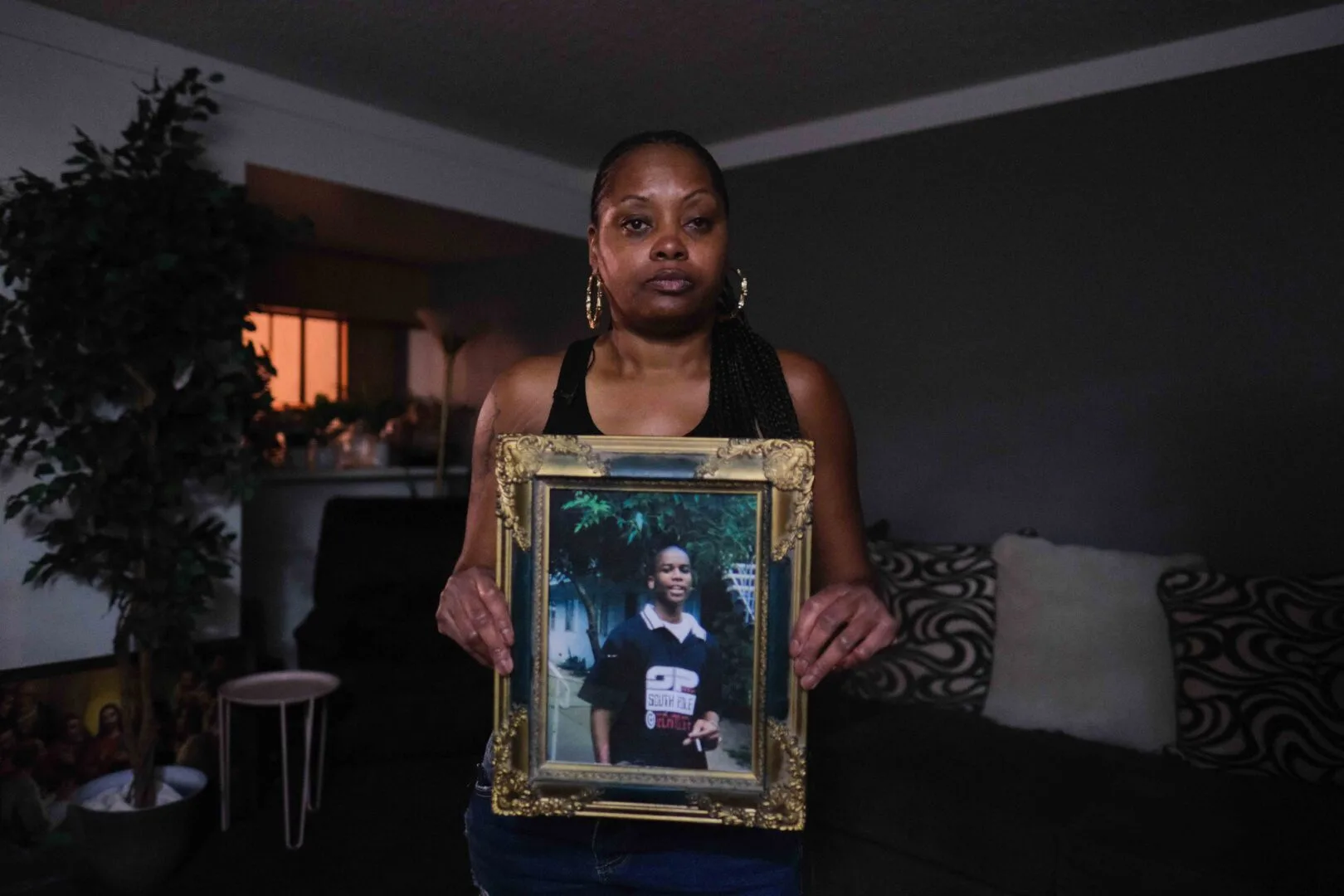 Keishia Brunston, a Black woman with long corn rows and wearing gold hoop earrings, holds a framed photo of Deondre Brunston in her living room. 