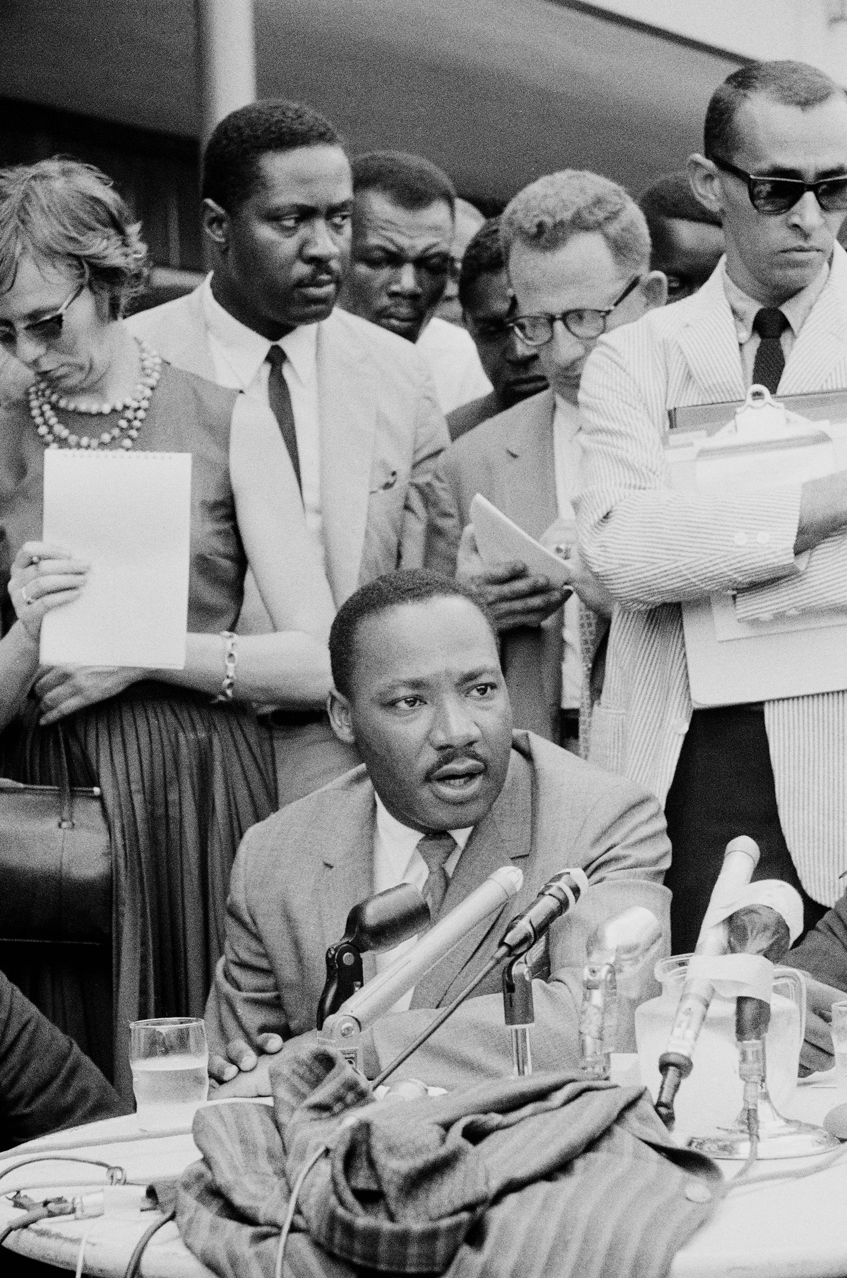 Dr. Martin Luther King Jr. sitting at a table behind microphones with a number of people behind him including Clarence...