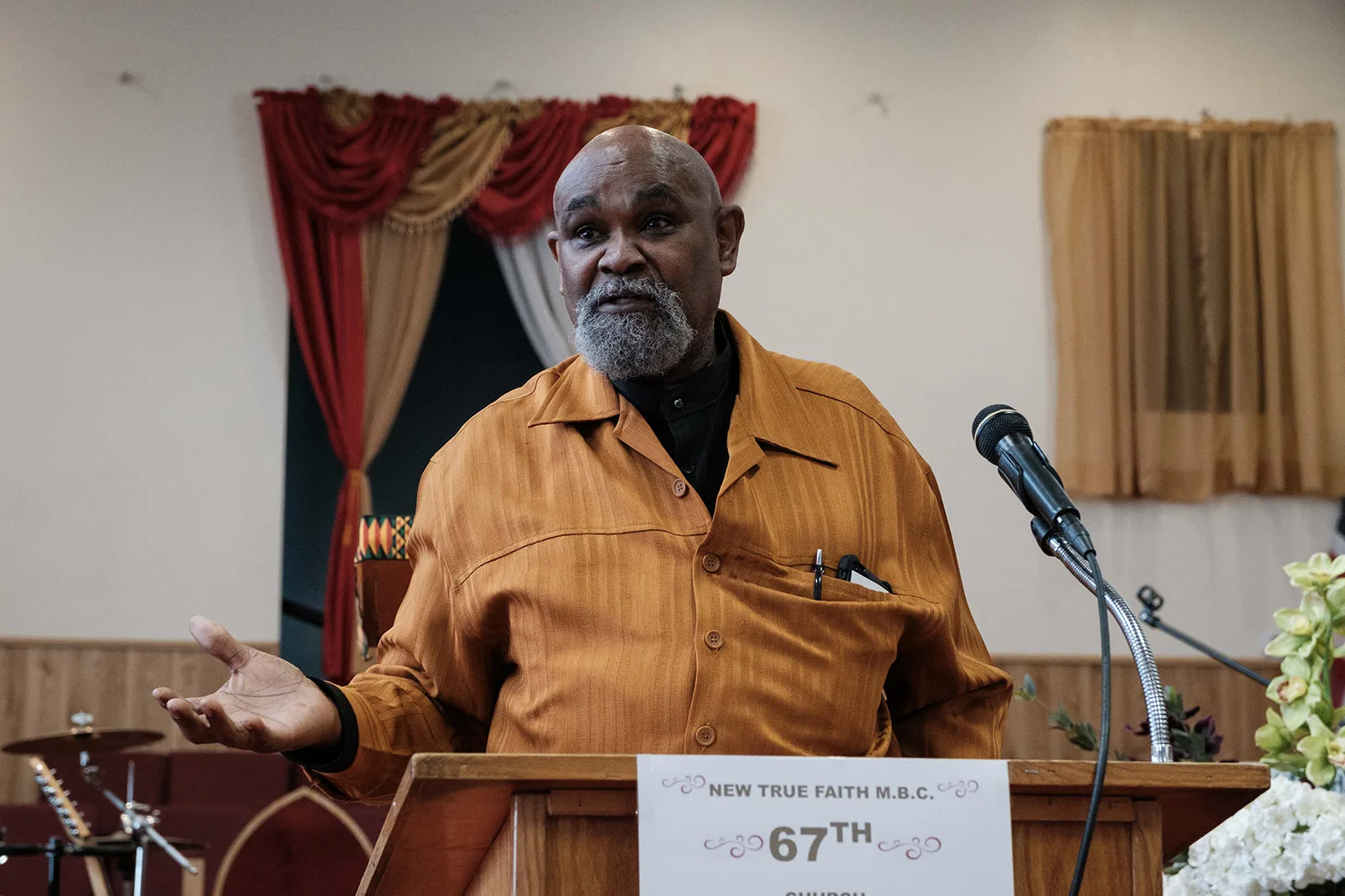 Pastor Thompson, A bald Black man with a grey beard wearing wearing a gold suit, stands at a podium and gestures to his left. 