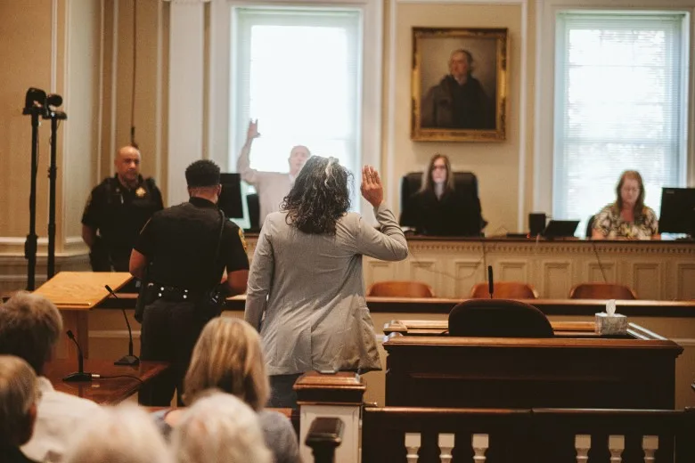 A view of a courtroom, with three people, including a judge, sitting at the dais in the background. Two uniformed officers move around in the middle ground. At the center of the photo, a woman in a pantsuit raises her right hand as people seated in the courtroom look on.