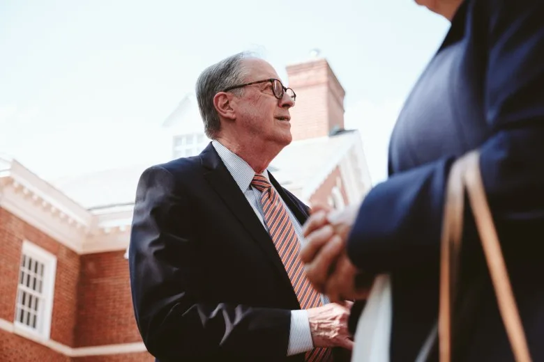 At the center of the photo, a man wearing glasses, a suit, and tie speaks to someone outside of the frame of the photo. In the background, a brick-and-white courthouse. In the foreground, someone walking into the frame.