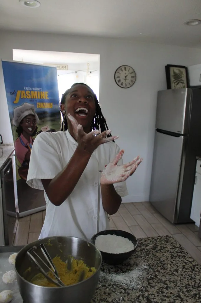 Kalea Barnes, a young Black woman, wears a white chef's jacket and stands in a residential kitchen. She laughs as she tosses flour into the air. In front of her on the counter is a large metal mixing bowl full of yellow cookie batter and a black bowl full of flour.