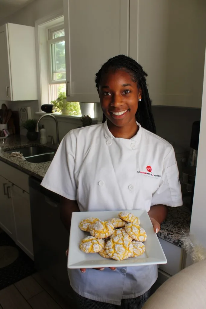 Kalea Barnes smiles while holding a square white plate stacked with white powder covered yellow cookies.
