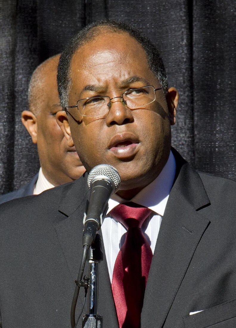 A man with brown skin and a close-cropped haircut graying at the temples stands in front of a microphone