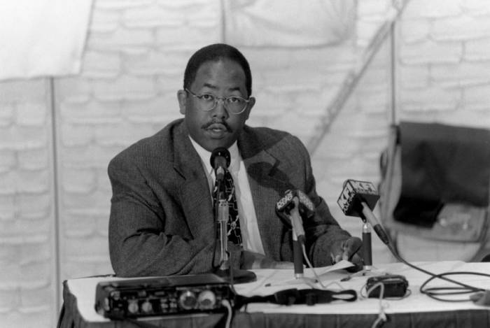 A black-and-white photo of young dark-skinned man with a close cropped haircut, wire-rimmed glasses and thin mustache sits at table in front of three microphones