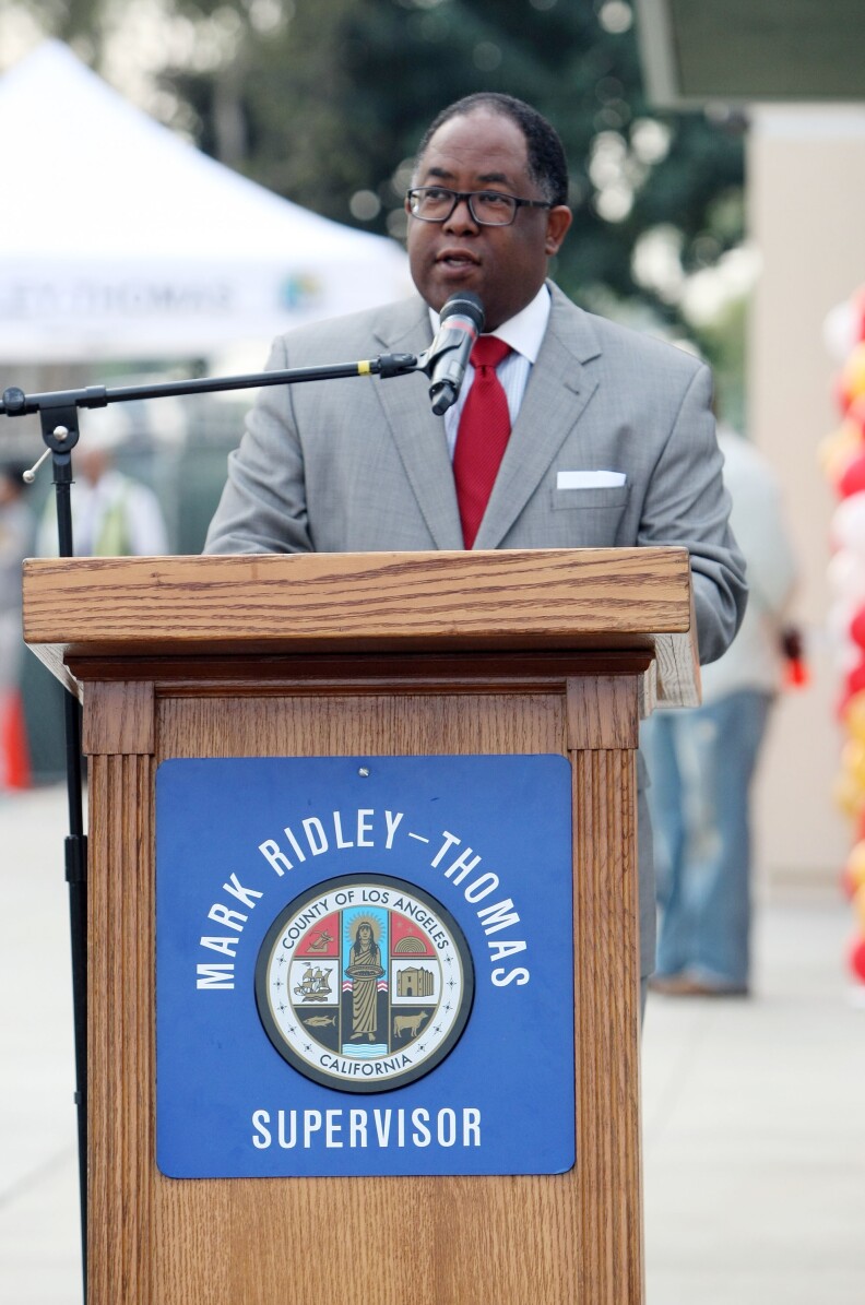 A middle-aged man with brown skin, a close-cropped graying haircut and wearing glasses, stands in front of a podium with a sign on the front that says Mark Ridley-Thomas, Supervisor, with an image of the seal of the county of Los Angeles
