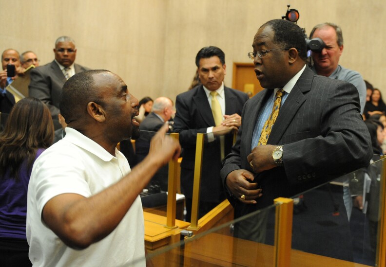 A brown-skinned man wearing a white polo shirt wags his finger at a brown-skinned man wearing a gray suit, blue shirt with a white collar and a patterned yellow tie at a council meeting. Several other people stand behind them in the council chamber.