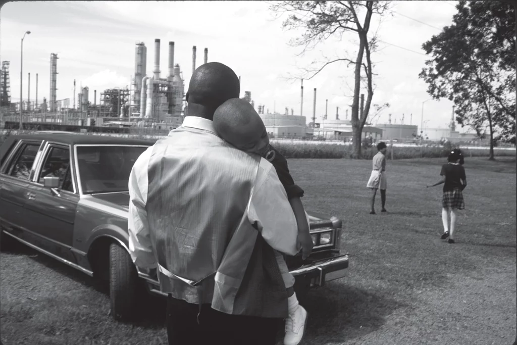 A family leaves Sunday church services in Lions, Louisiana, in October 1998. In the foreground, a man holds a sleeping child on his shoulder, standing in front of a car, with two women visible in the midground. An industrial chemical plant looms in the background. 