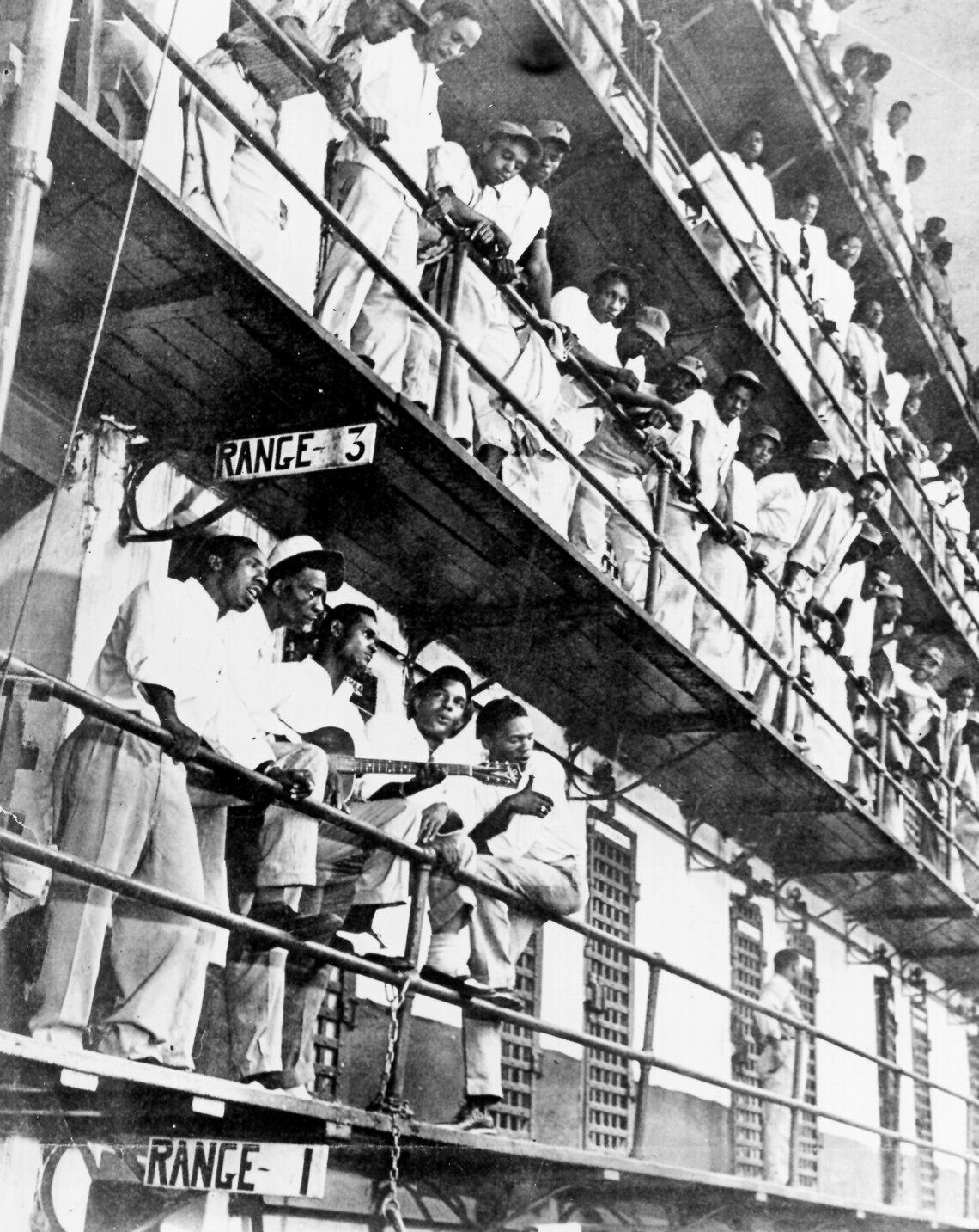 Five members of the singing group The Prisonaires lean against the railing of a balcony in a prison hall. One of the musicians is holding a guitar while the other men sing around him. On increasingly higher balconies above the group, prisoners peer over the edge of the railings, watching the performance.