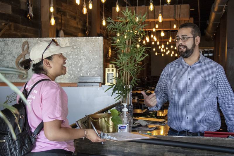 Two people talk inside of a restaurant. One person is holding a clipboard.