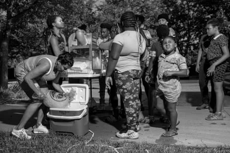 Children standing around a snowcone machine, a woman adds ice to a cooler next to them.