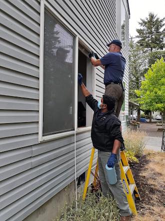 Standing outside a grey-sided house, one person stands with a hand toward a window next to another person on a ladder, working slightly above the first person. Both are doing work for Taking Ownership PDX, a Portland organization that treats Black home ownership as a social justice issue.