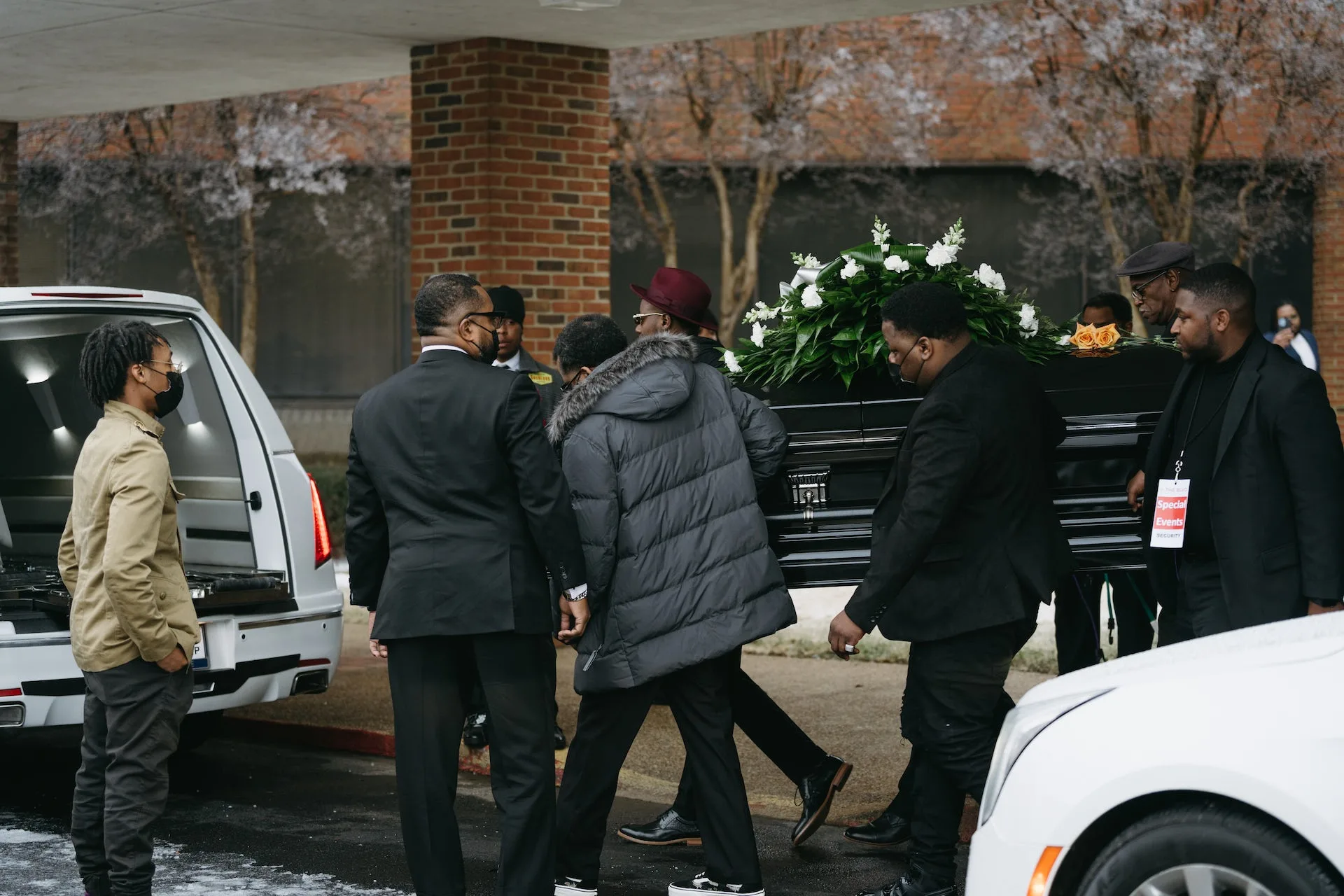 Mean wearing black and gray carry a black casket topped with white flowers to the open back of a white hearse.