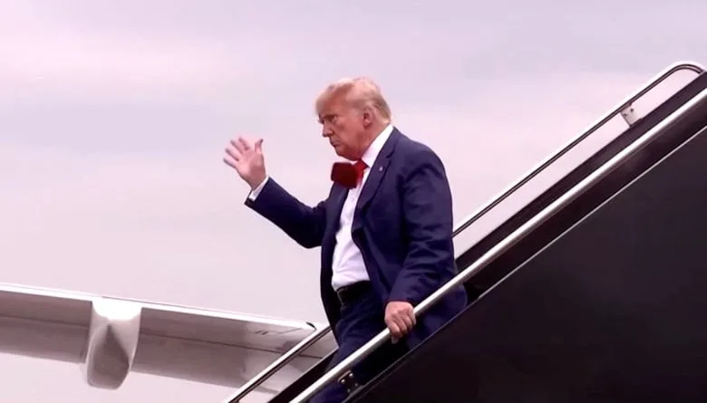 Trump walks down plane stairs, his tie flapping in the wind