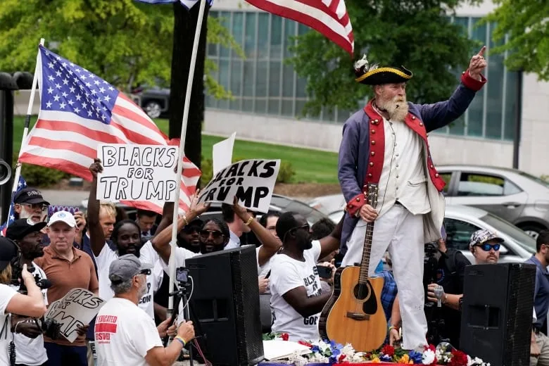 Man dressed as 18th century revolutionary with a guitar in front of men holding signs that say 'Blacks For Trump' 