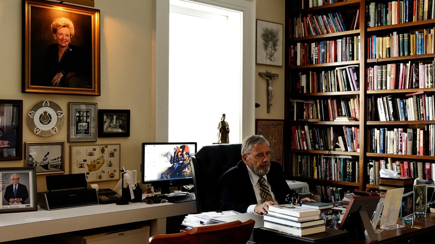 A man in a suit sits at a desk with a computer and a bookshelf in the background.