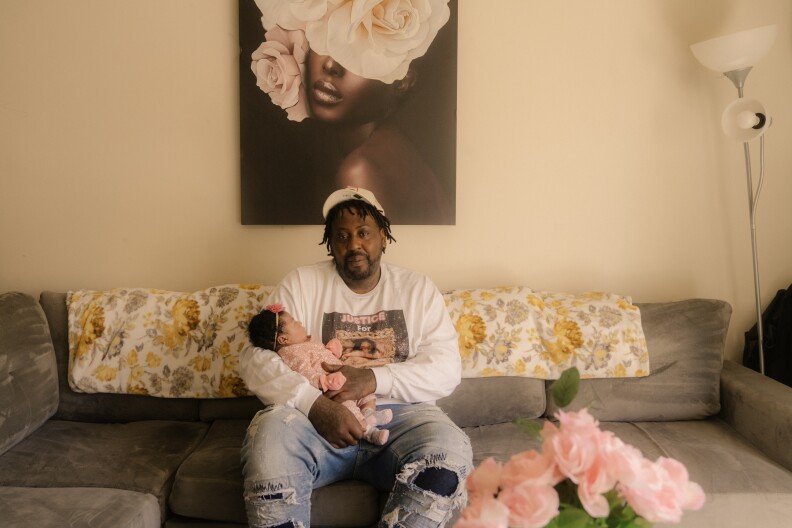 A Black man wearing a white baseball hat, long sleeve white t-shirt and jeans sits on a gray couch while holding a small baby wearing a pink onesie and a headband with a pink flower. Behind him there is a print hanging on the wall of a Black woman with flowers on her face. In the foreground to the left of the man's leg, there are pink flowers. 