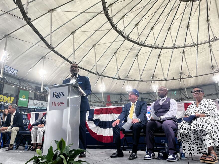 Man in a suit stands at a podium inside a domed stadium. Other people sit to his left and right. 