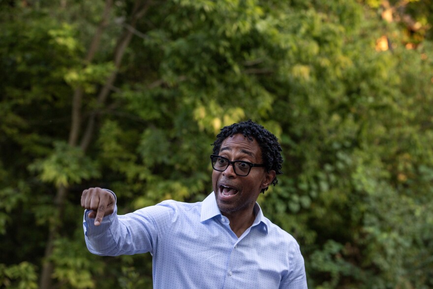 Missouri politician Wesley Bell uses a downward pointed finger to make a point while speaking. Behind him is a lush scene of greenery and trees. 