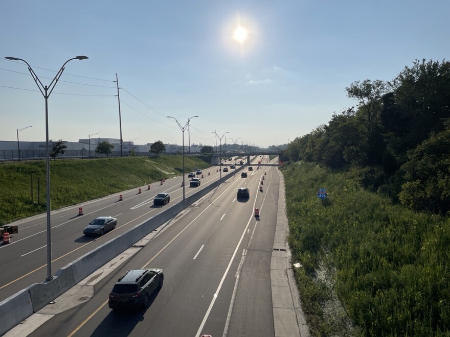 An image of Westbound I-496 in the afternoon, with the sun centered in a blue sky and several cars driving down the freeway alongside traffic cones.