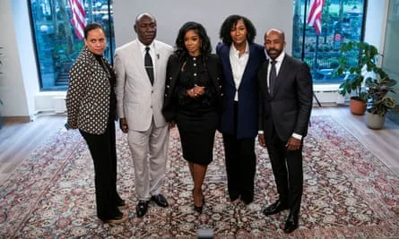 A line of five Black adults, three women and two men, dressed in suits pose for a picture on a rug in an office building.