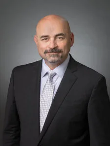 This is a photo of Joshua Malcom, a member of the lottery commission. He's a middle-aged Native American man with very short hair, a brown-gray mustache and beard. He's wearing a black suit jacket and silver tie and white shirt.