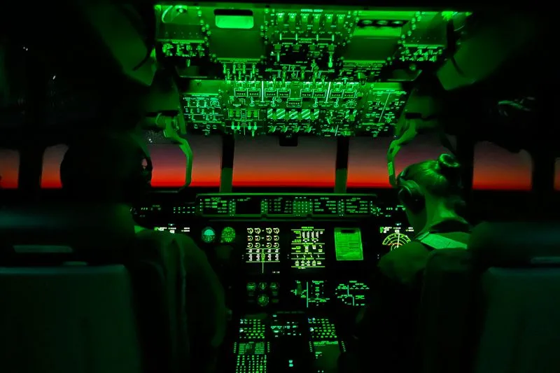 A view from the cockpit shows backlit control panels and two pilots inside a KC-130J aerial refueler en route from Williamtown to Darwin as the sun sets on the horizon.