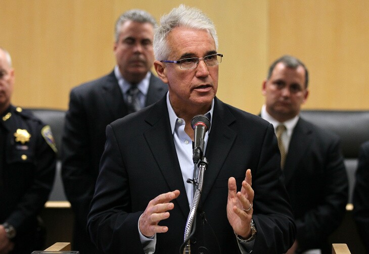 L.A. district attorney George Gascón, a man with light skin tone, grey hair and glasses, is speaking at a podium. Officials are behind him.