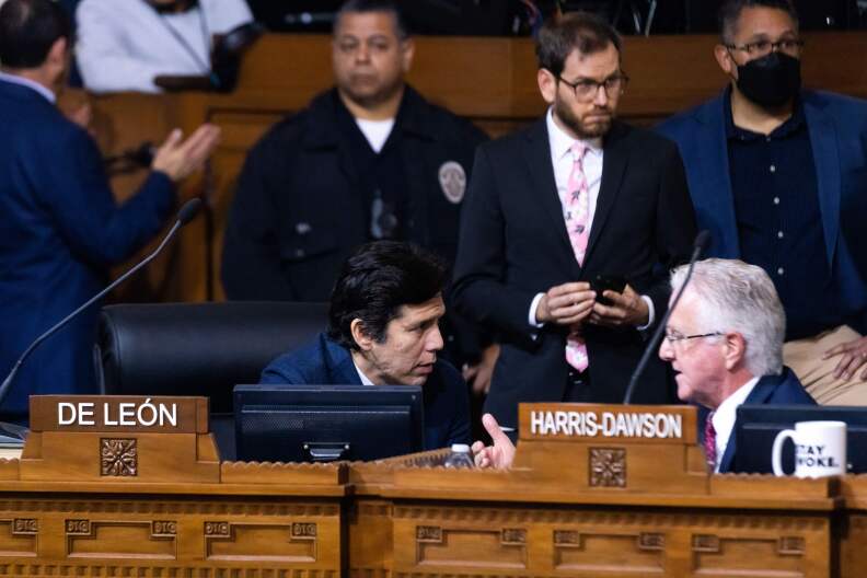 Kevin de León sits behind his wooden dais he turns towards his left as he speaks to Paul Krekorian, who is in Marqueece Harris-Dawson's seat.  There are three men standing behind them. 