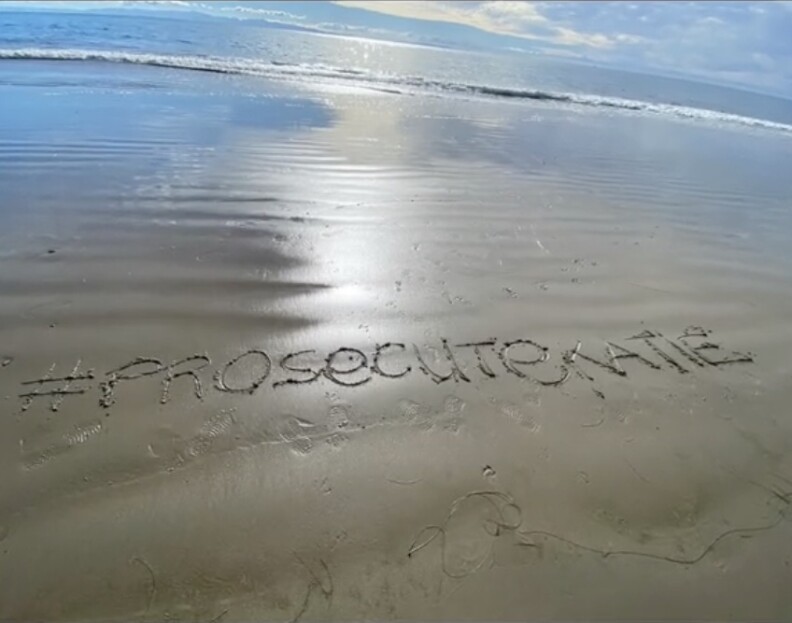 The words #ProsecuteKatie are written in wet sand near the shoreline