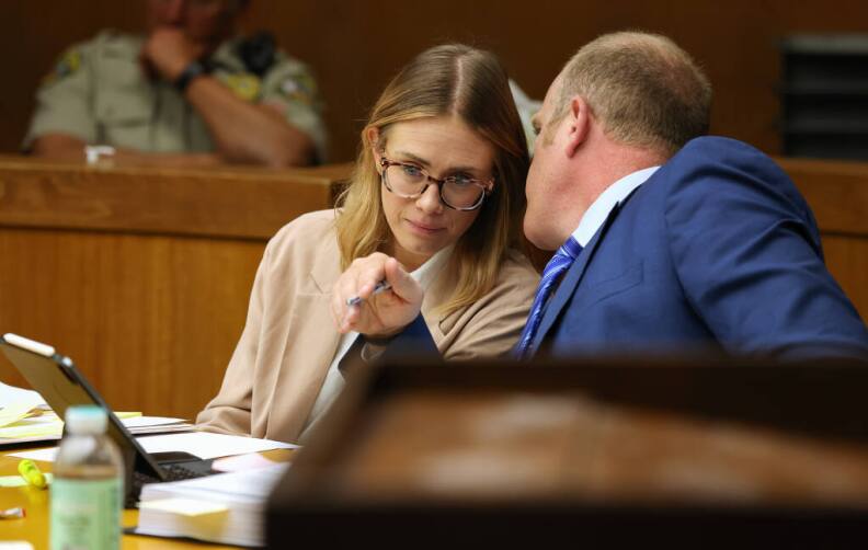 A white woman in a taupe jacket leans over as a man in a dark blue suit speak into her ear. they're at a table in a court.