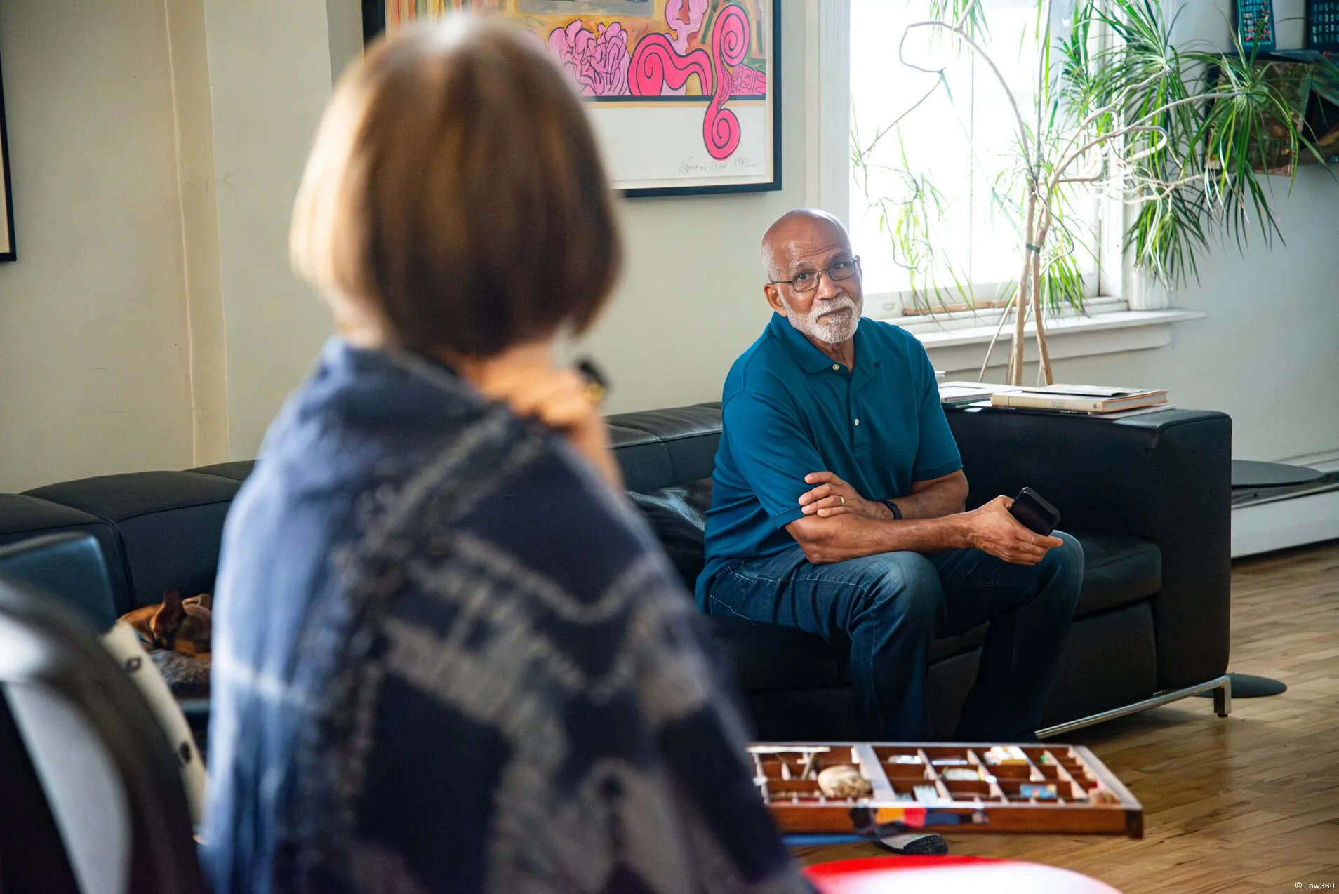 A middle aged white woman and a middle aged Black Hispanic man chat while sitting on a sectional sofa. The photo is taken from over the woman's shoulder, and is focused on the man.