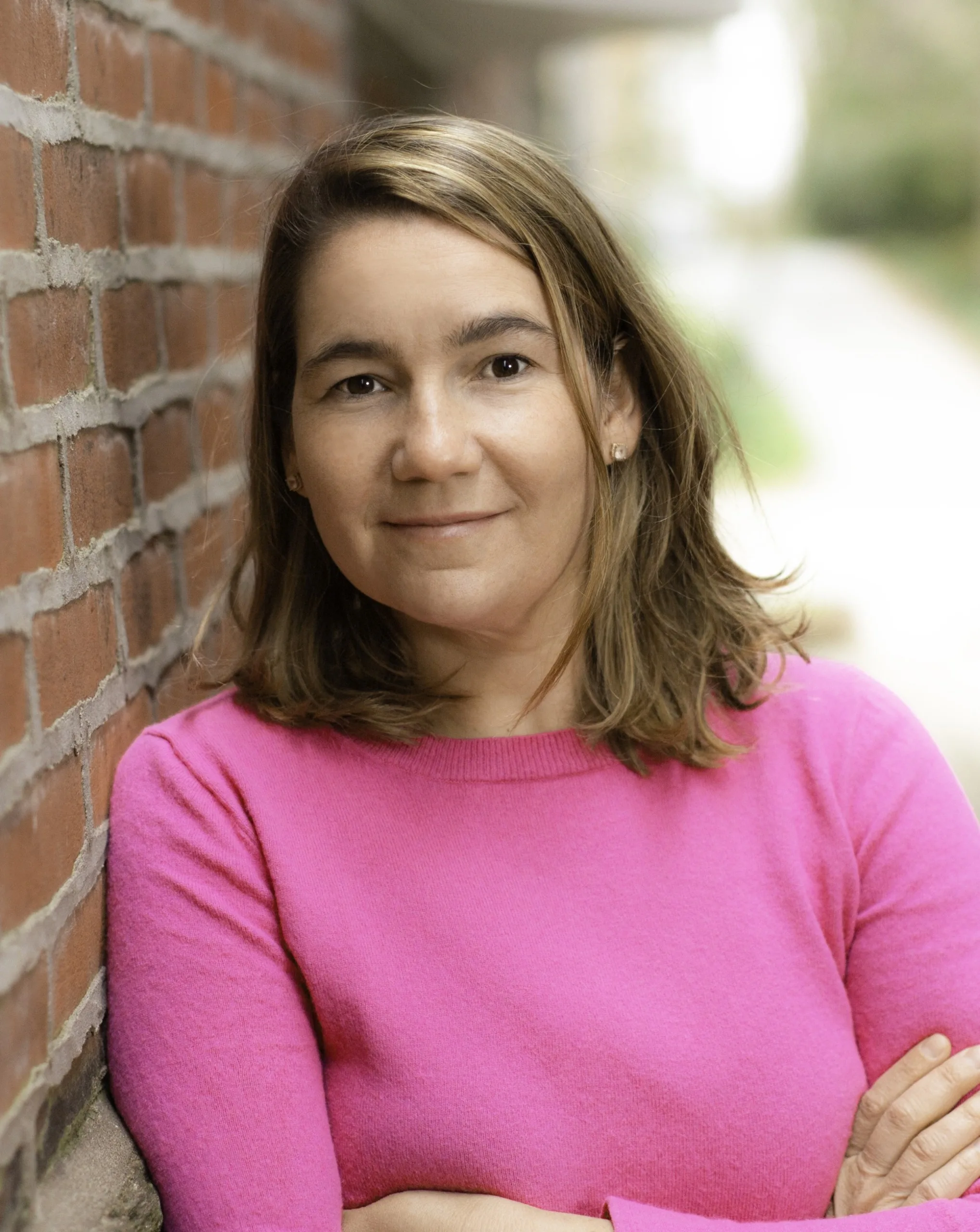 Natalie Renew headshot: white woman in pink shirt leaning against brick wall