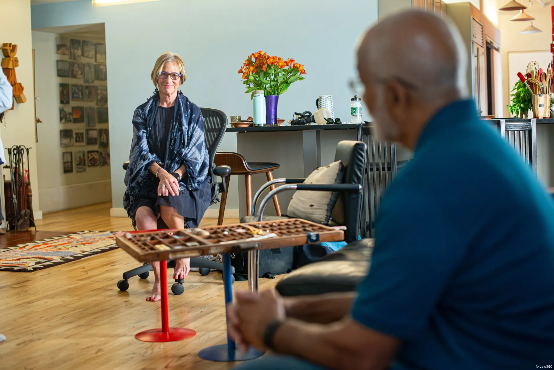 A middle aged white woman and a middle aged Black Hispanic man chat while sitting on a sectional sofa. The photo is taken from a perspective just behind the man, and is focused on the woman.