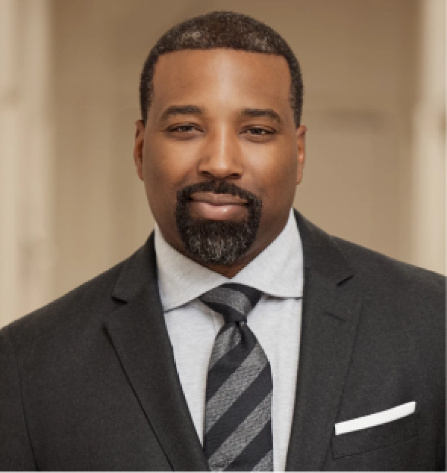 A headshot of Michael Phillips facing the camera. He is in a black suit, white undershirt, and black tie.