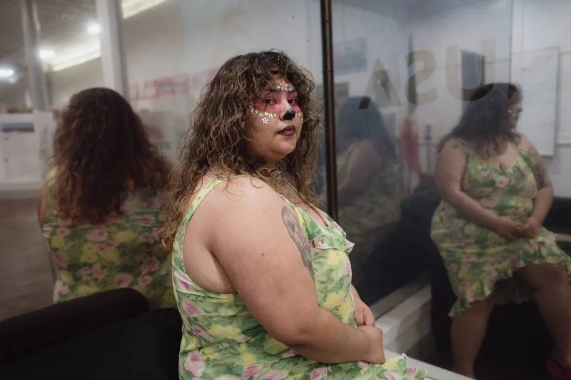 A woman wearing face paint and a green, floral print dress stands next to a window.