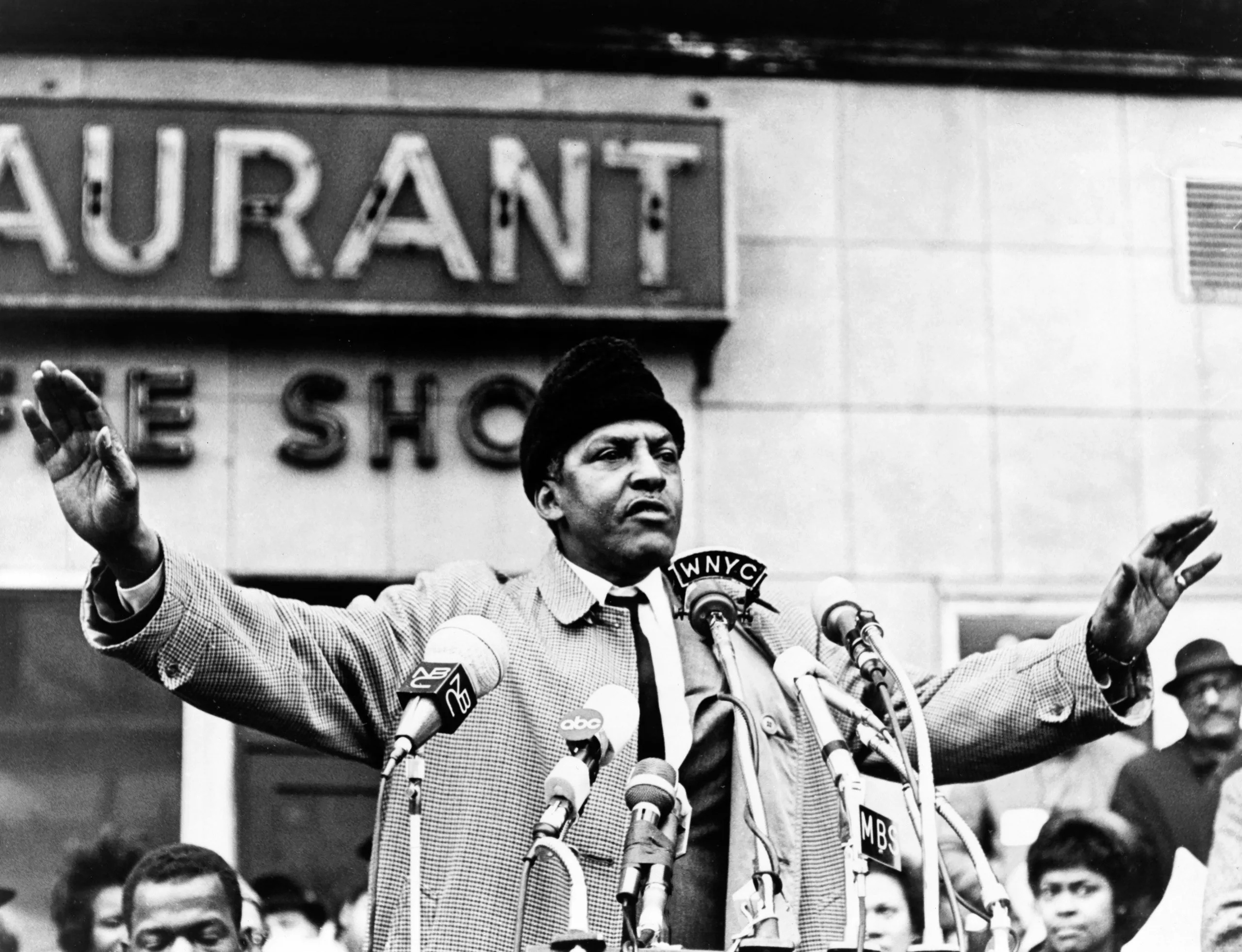 Bayard Rustin speaks before a number of microphones with his hands raised in the air.