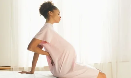 Stock image of young Black pregnant woman in pink gown sitting on edge of examining table and holding back with one hand.