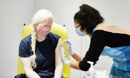 Latina woman in face mack injects shoulder of albino woman with long pale braid.