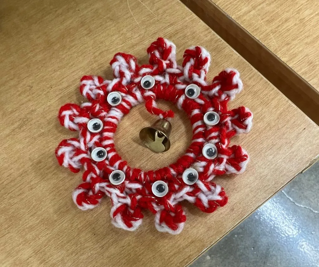 A handmade ornament with red and white yarn and google eyes with a bell in the center.