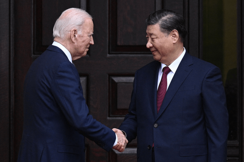 U.S. President Joe Biden is seen in profile as he greets Chinese President Xi Jinping with a handshake. Xi, a 70-year-old man in a dark blue suit, smiles as he takes the hand of Biden, an 80-year-old man who also wears a dark blue suit.